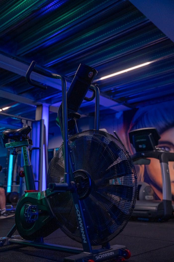 A modern gym filled with exercise equipment under blue lighting, featuring a stationary fan bike.